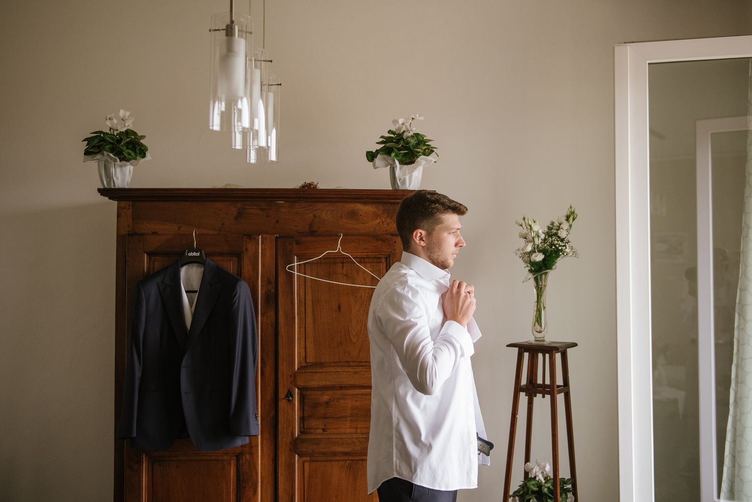 Groom preparing in a stylish room with a wooden wardrobe and his suit hanging nearby.