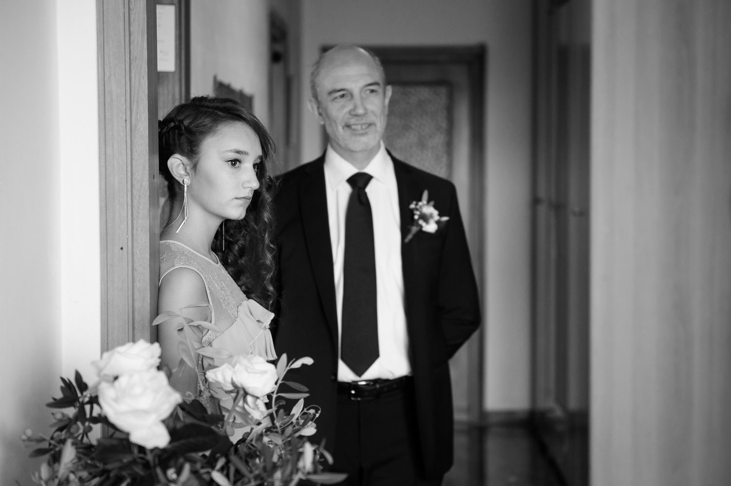 Black and white photo of a bride holding flowers standing with an older man in a suit indoors.