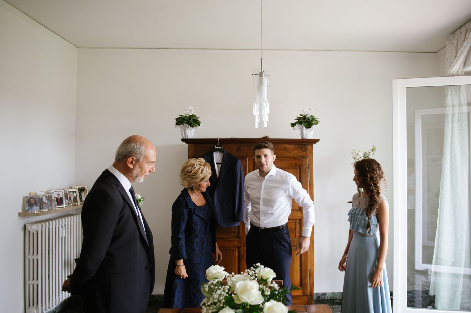 Groom in white shirt stands with family members in elegant room with floral arrangement before wedding ceremony.