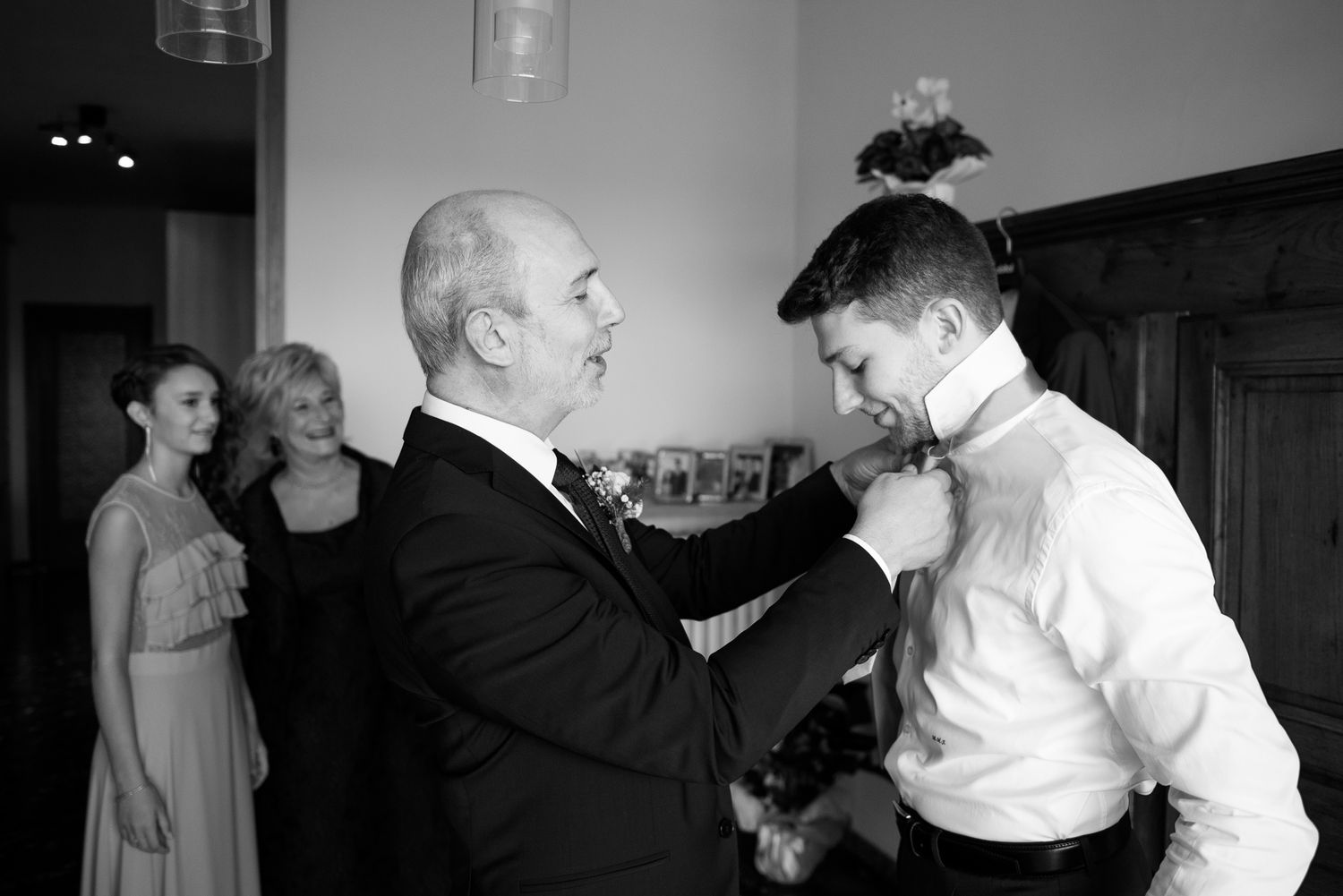 Older man adjusting grooms tie while bridesmaids watch during wedding day preparations indoors.