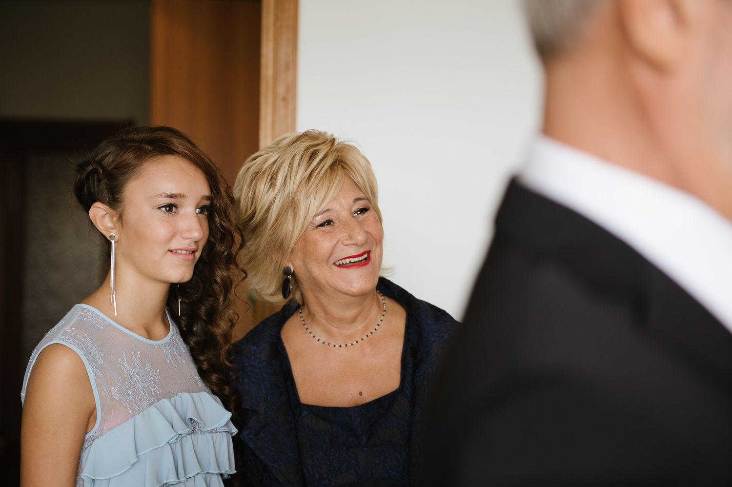 Young woman and older woman in formal attire smiling warmly at a wedding celebration indoors.