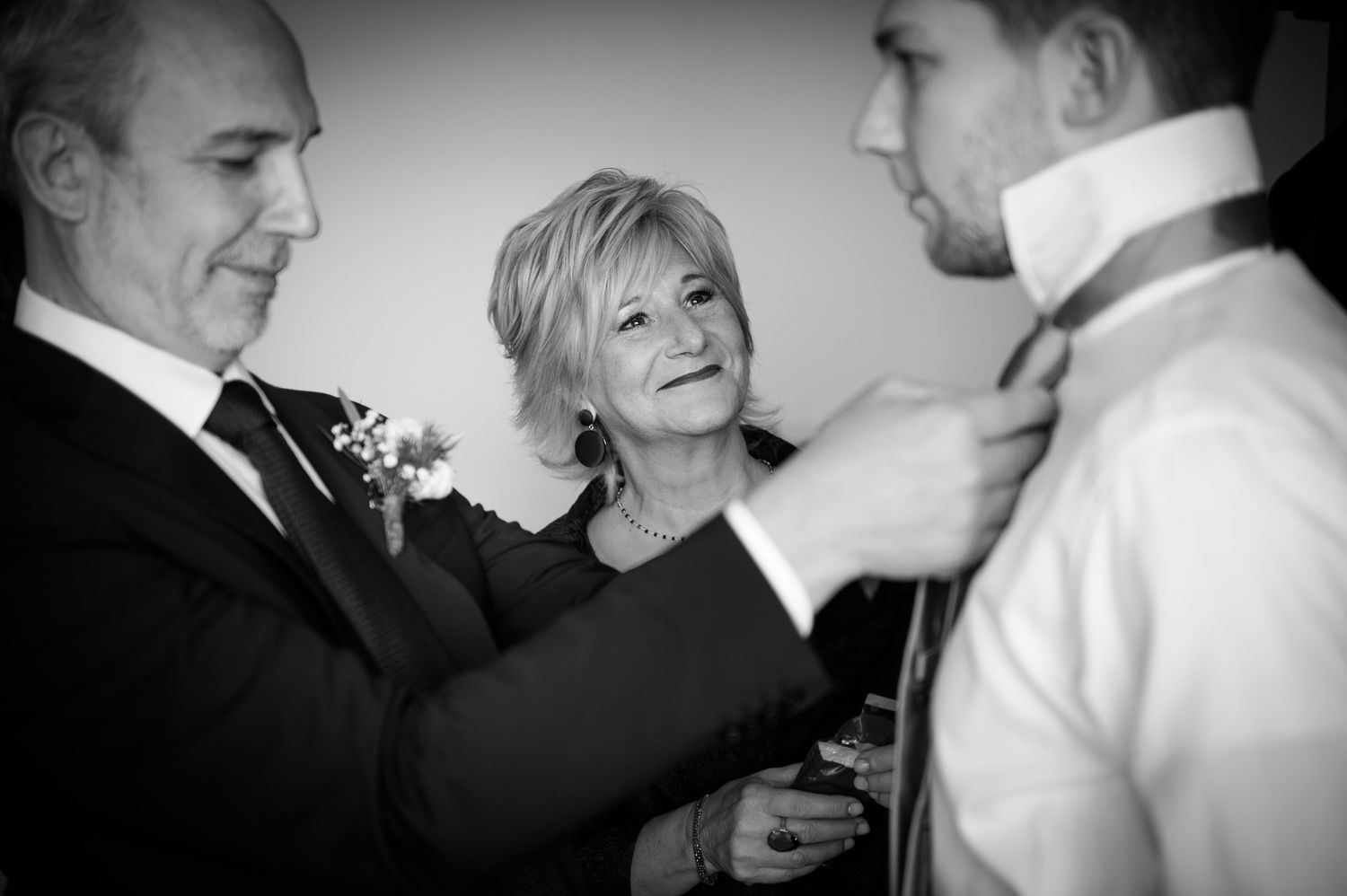 Parents helping groom adjust his tie before wedding ceremony in black and white photo.