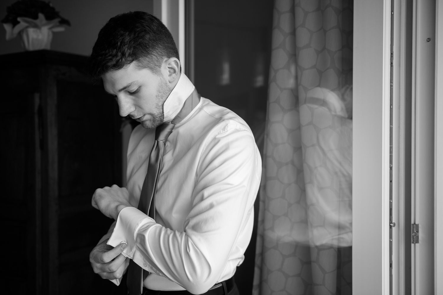 Black and white photo of a groom adjusting his cufflinks by a window.