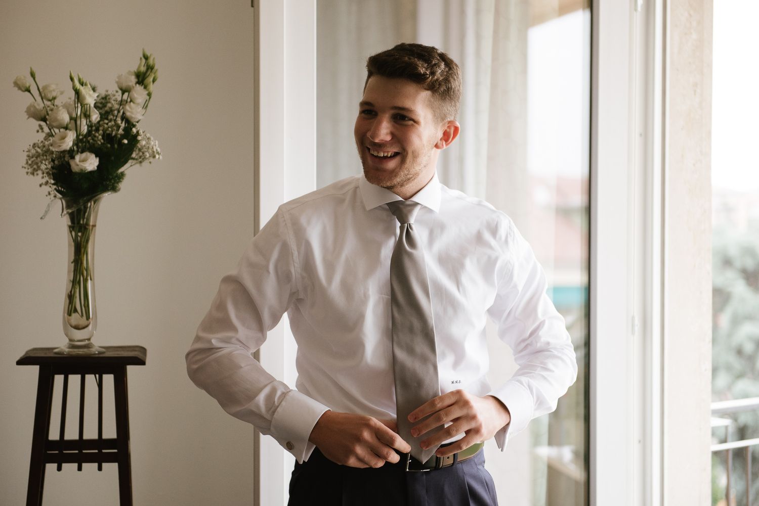 Smiling groom in white dress shirt and tie buttoning jacket near bright window on wedding day.