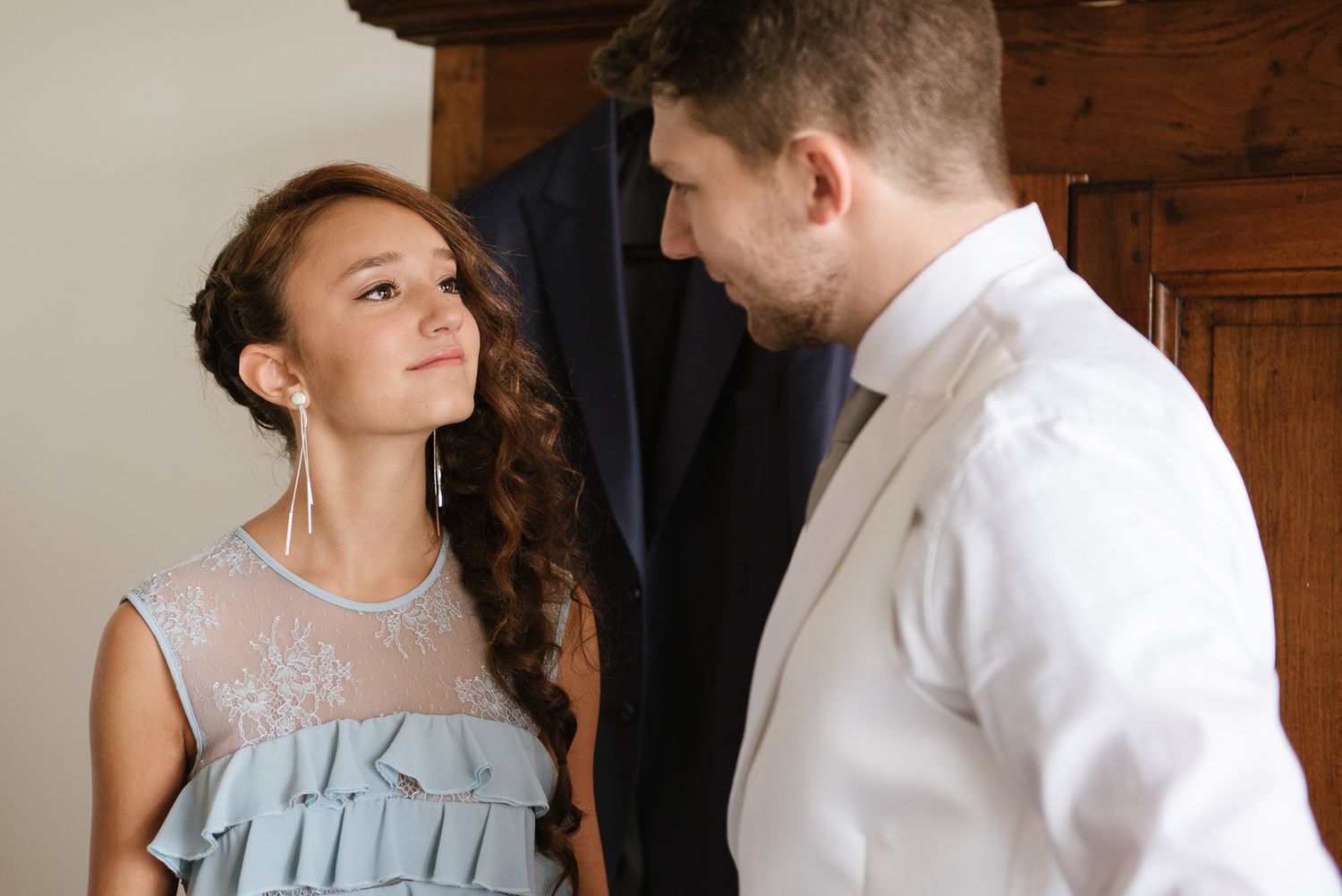 Young bridesmaid in blue dress gazing up at groom in white suit jacket before wedding.