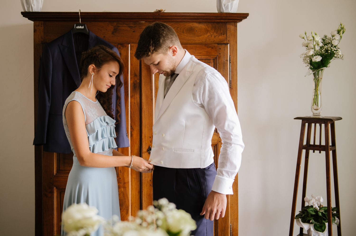 Bride in blue dress helping groom button his white shirt near wooden wardrobe before wedding ceremony.