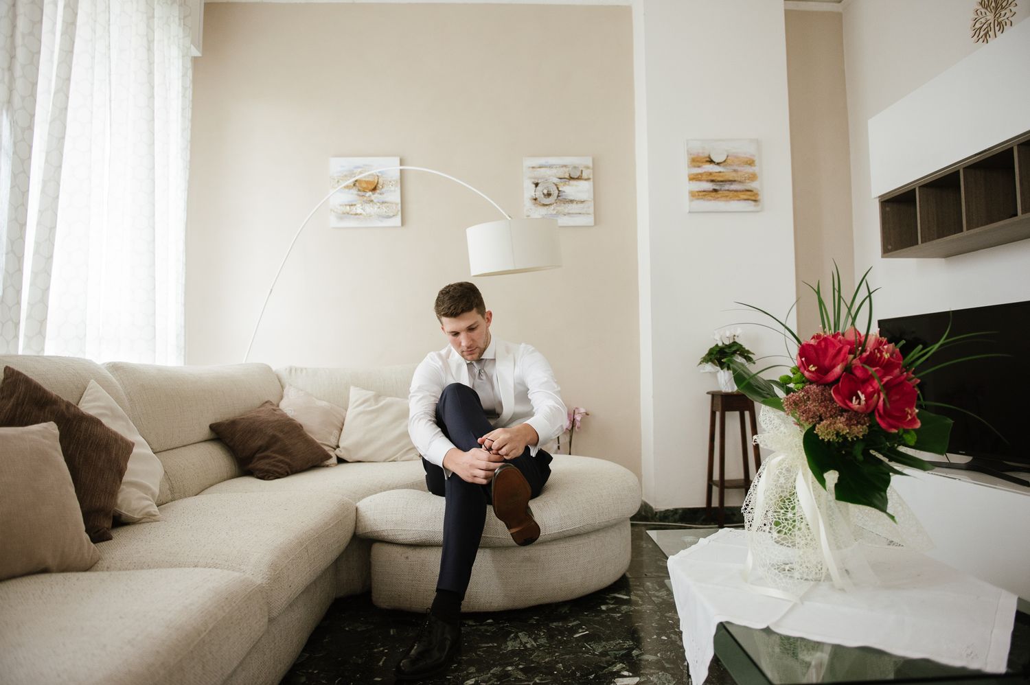 Groom in white shirt sitting on beige sofa putting on shoes in elegant living room before wedding.