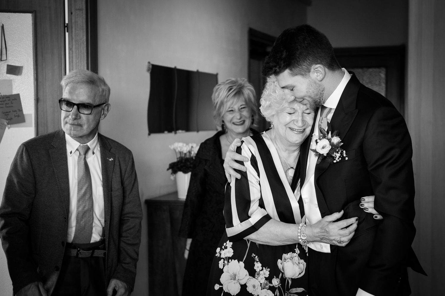 Groom tenderly embraces elderly woman while family members smile during emotional black and white wedding preparation photo.