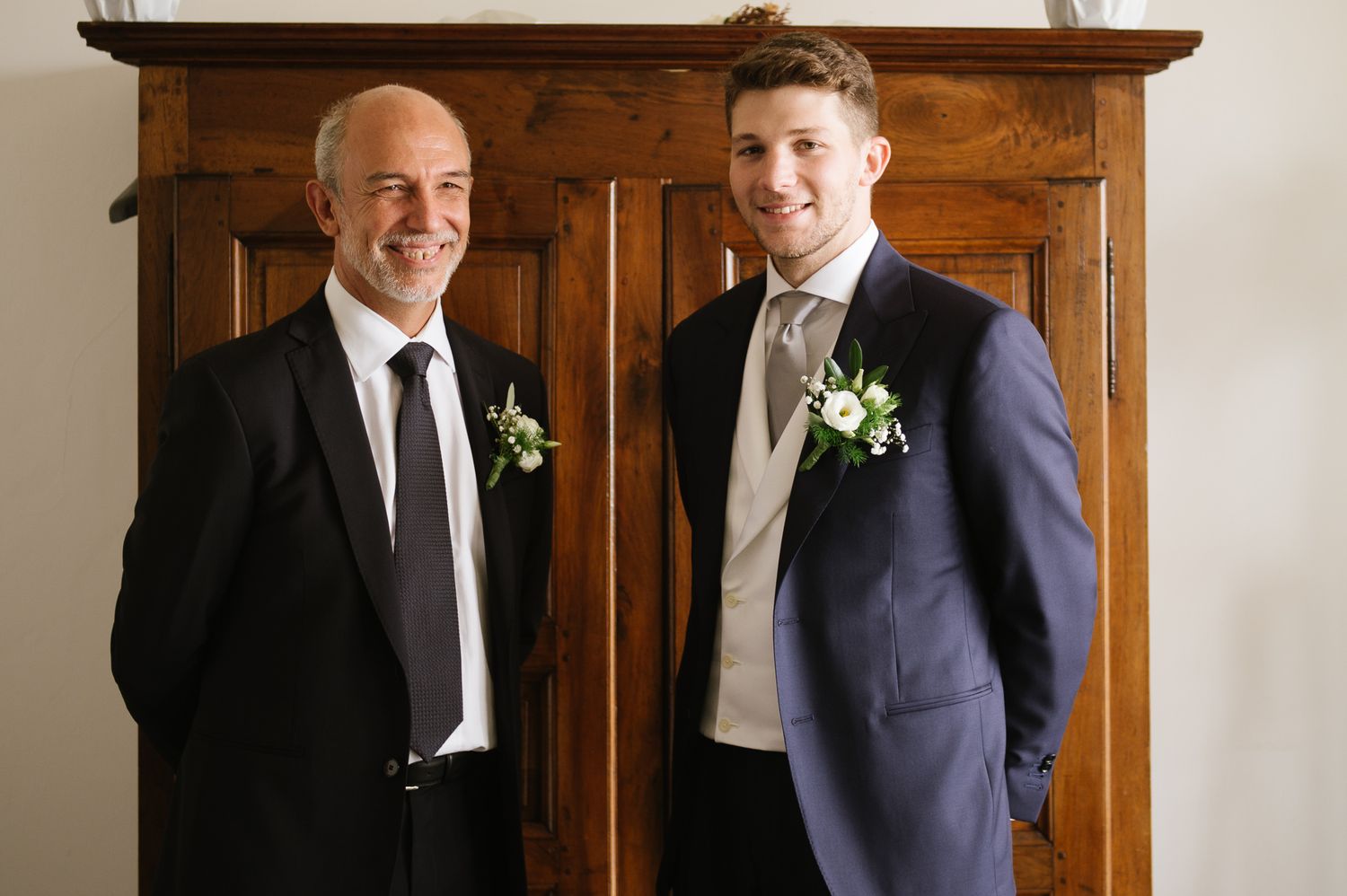 Two men in suits with white floral boutonnieres smiling together in front of a wooden armoire.