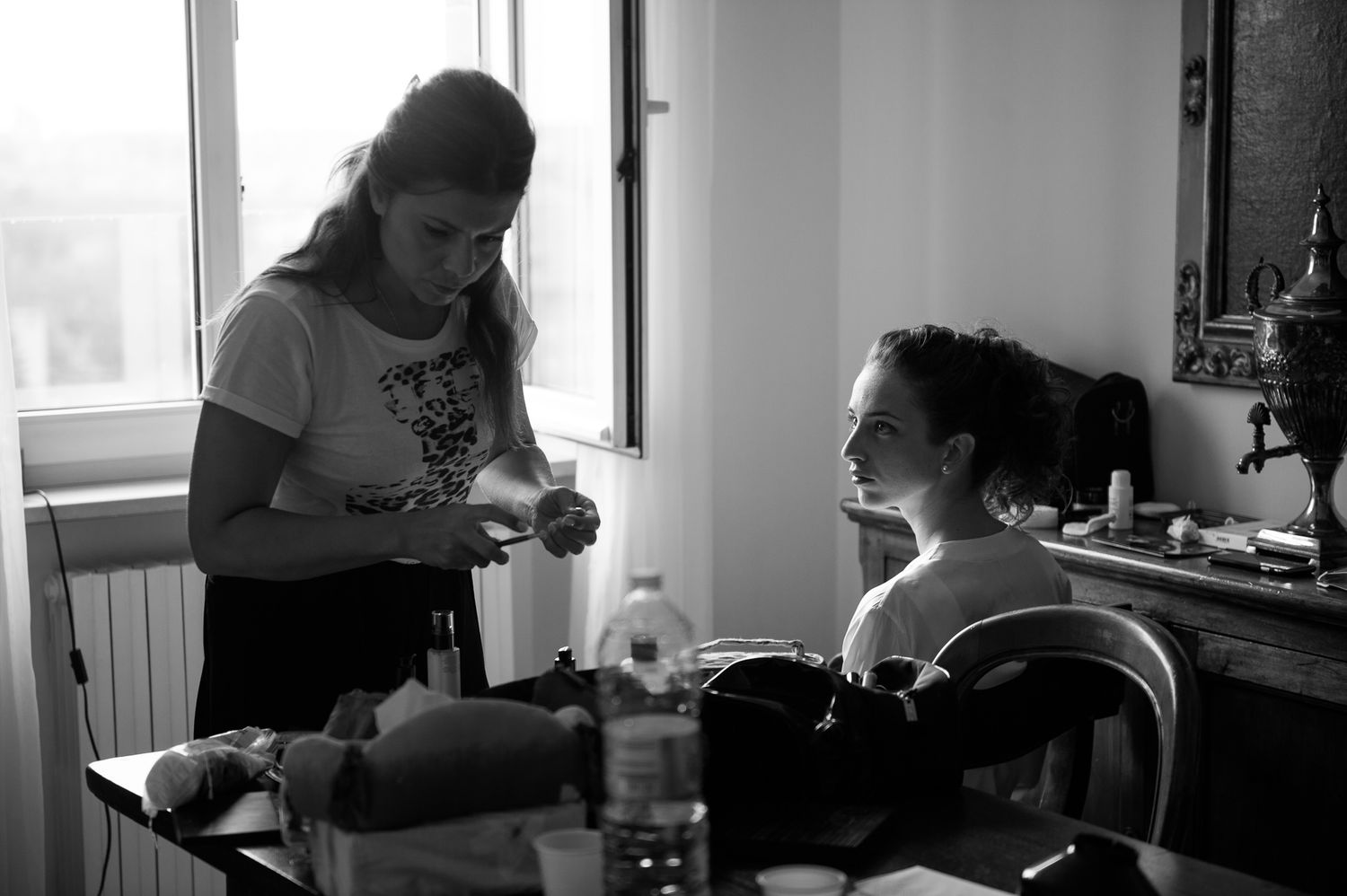 Makeup artist preparing bride for wedding day in bright room with beauty products on table.