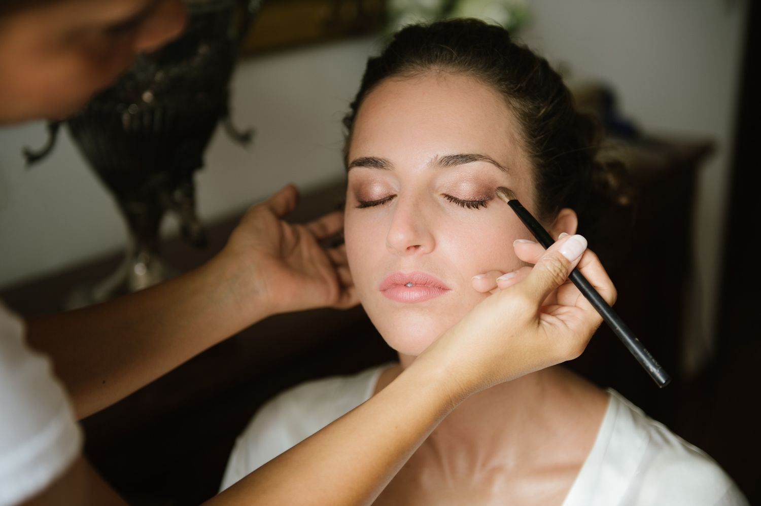 Professional makeup artist applying eye makeup to brides closed eyes before wedding ceremony.