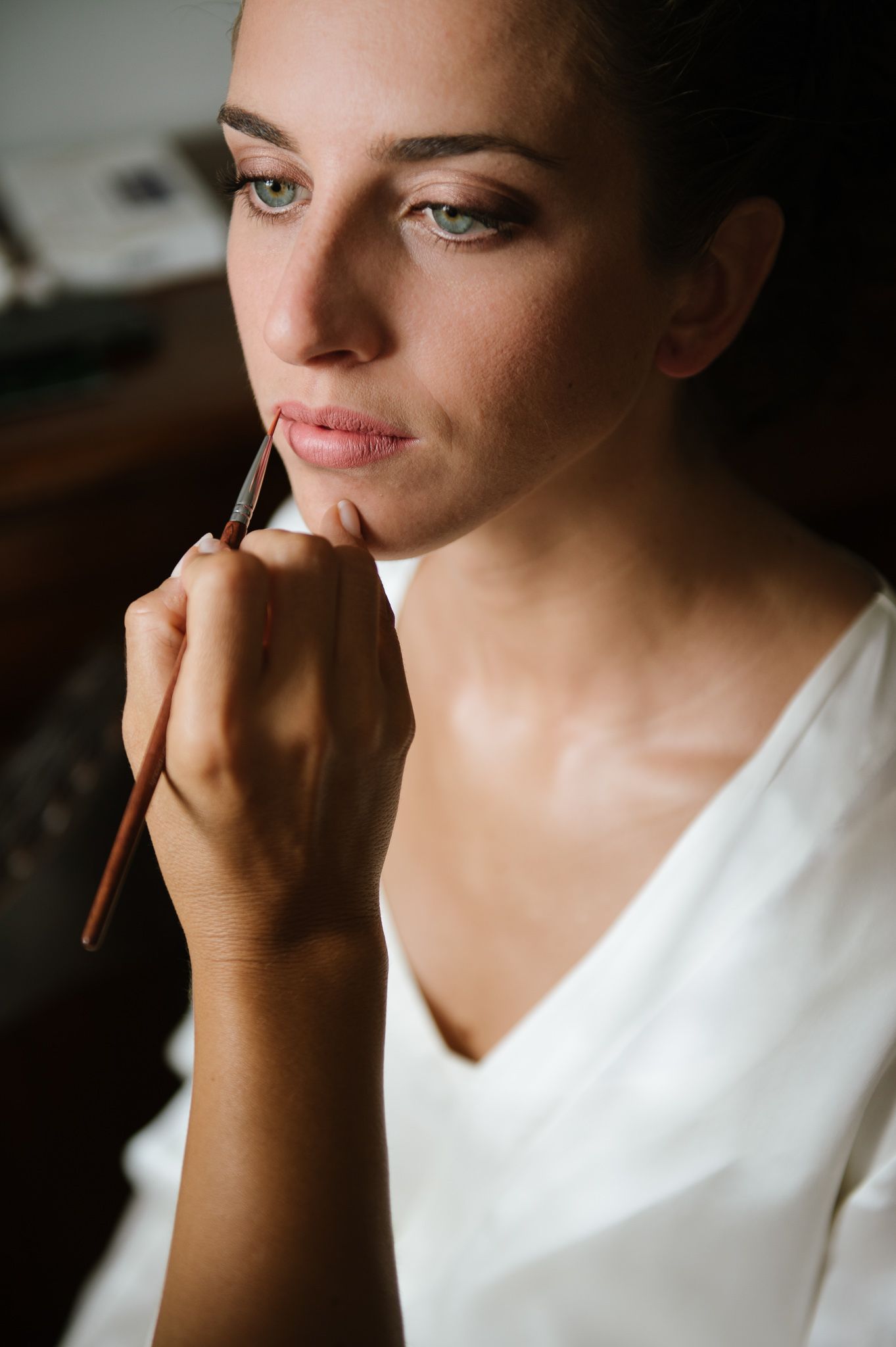 Bride applying lip makeup with brush during wedding preparation in natural light.