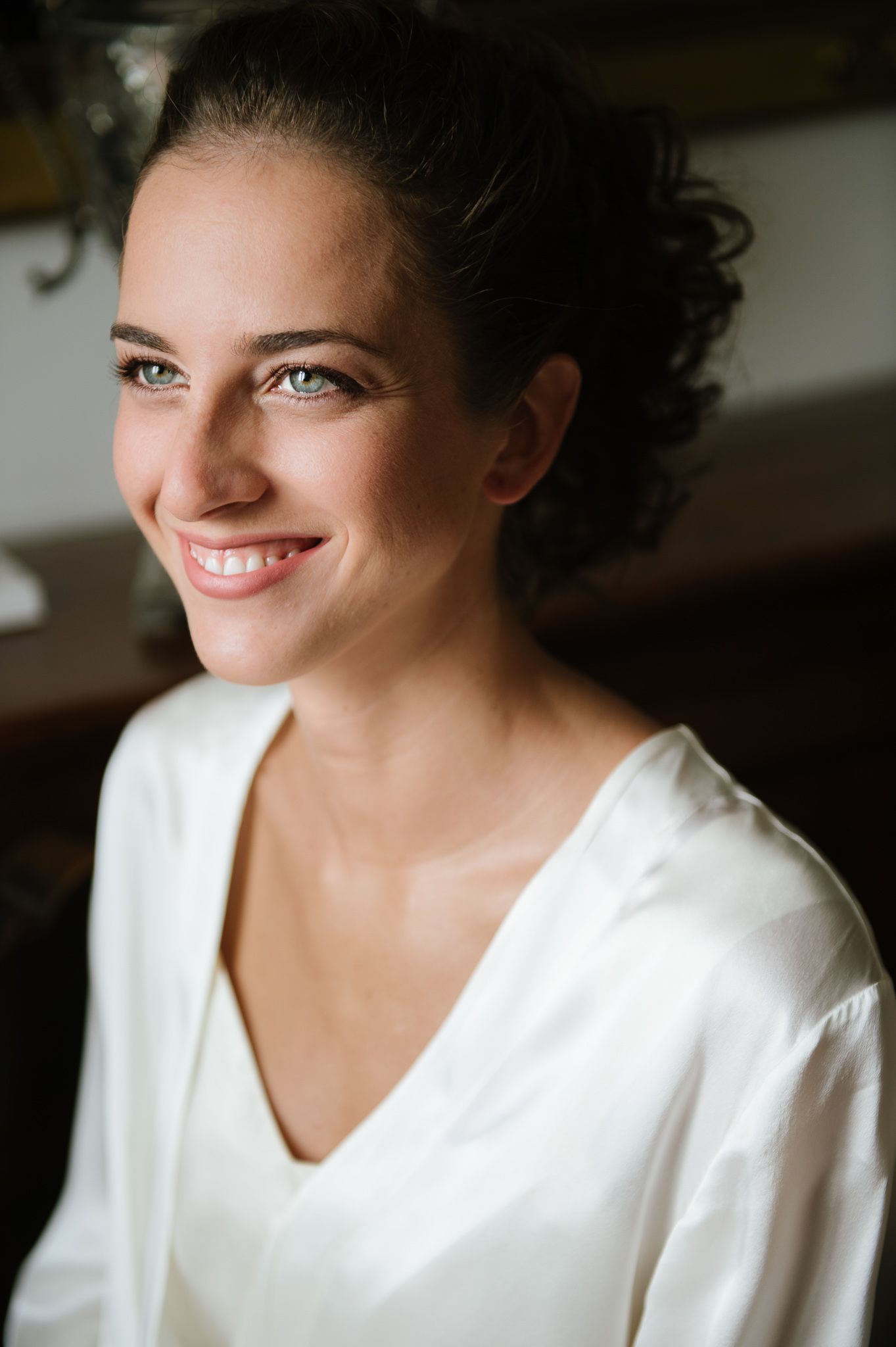 Beautiful bride with blue eyes and dark hair in an updo, wearing a white dress, smiling softly before her wedding.