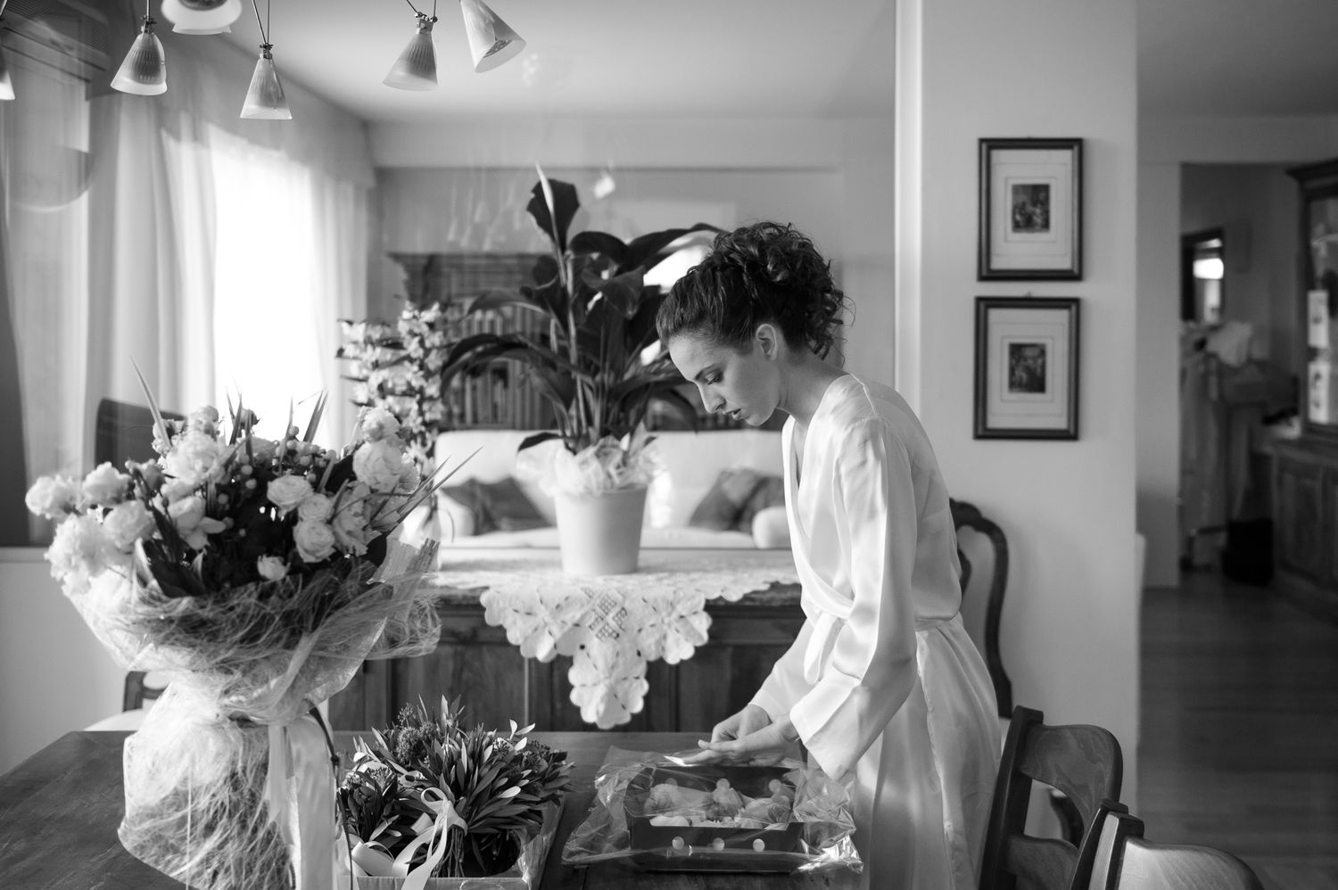 Black and white photo of bride in robe arranging items on a table surrounded by wedding flowers.