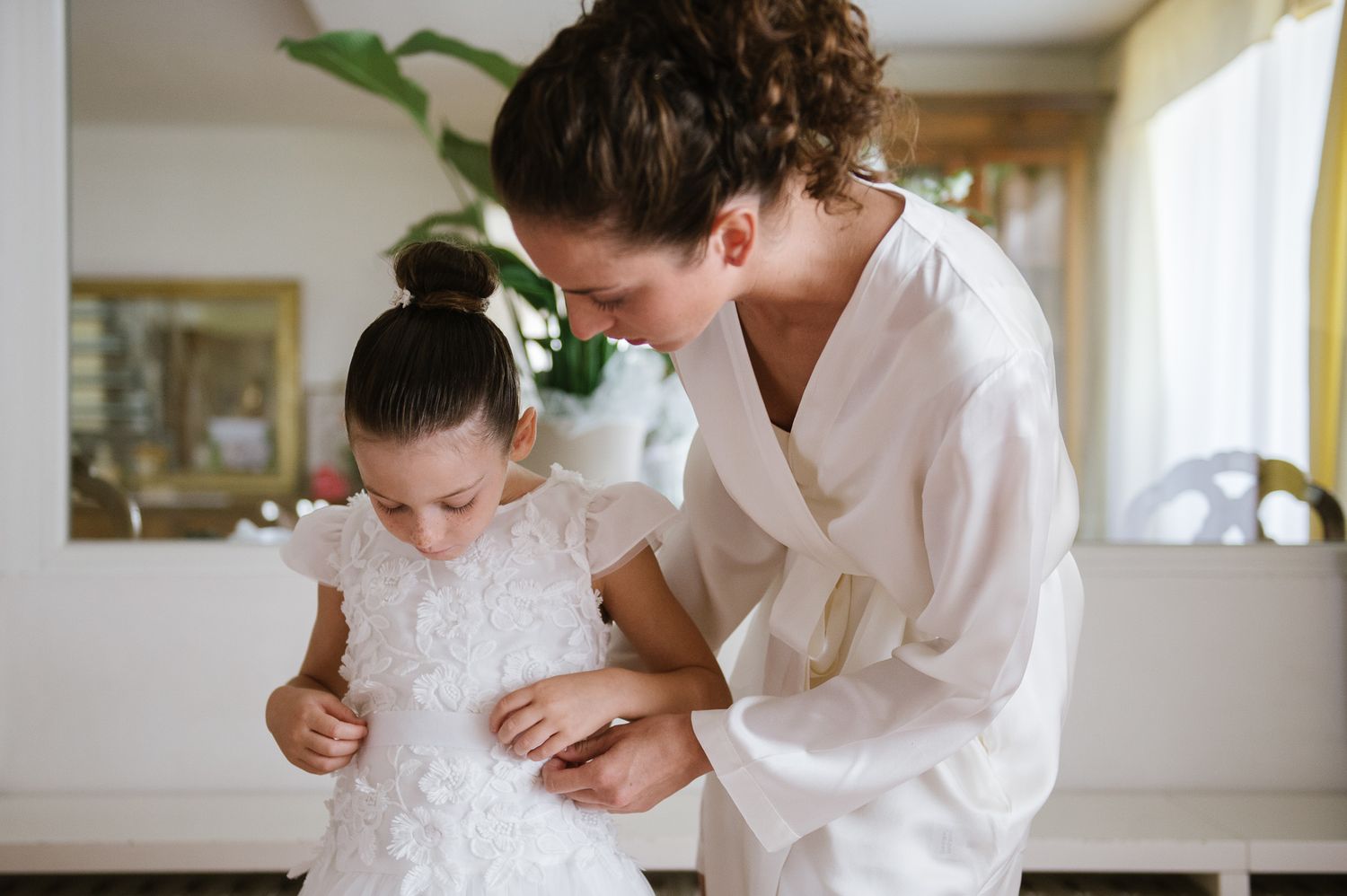 Bride in white robe carefully adjusts flower girl dress during intimate wedding morning preparation moment.