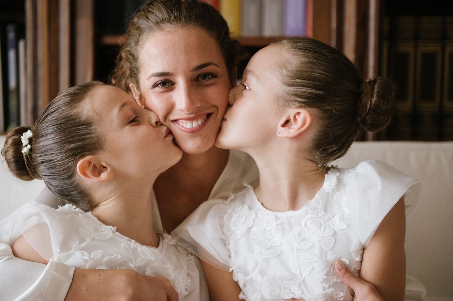 Smiling bride in white dress receives kisses on cheeks from two young flower girls in white dresses.
