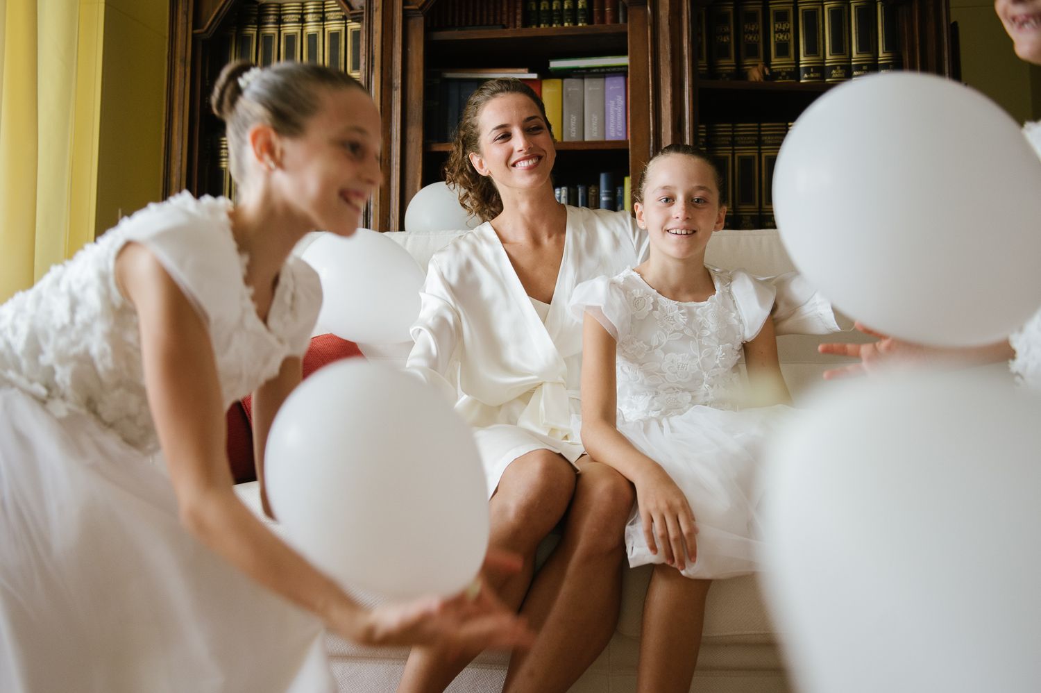 Bride and bridesmaids laughing together holding white balloons during wedding preparations indoors.