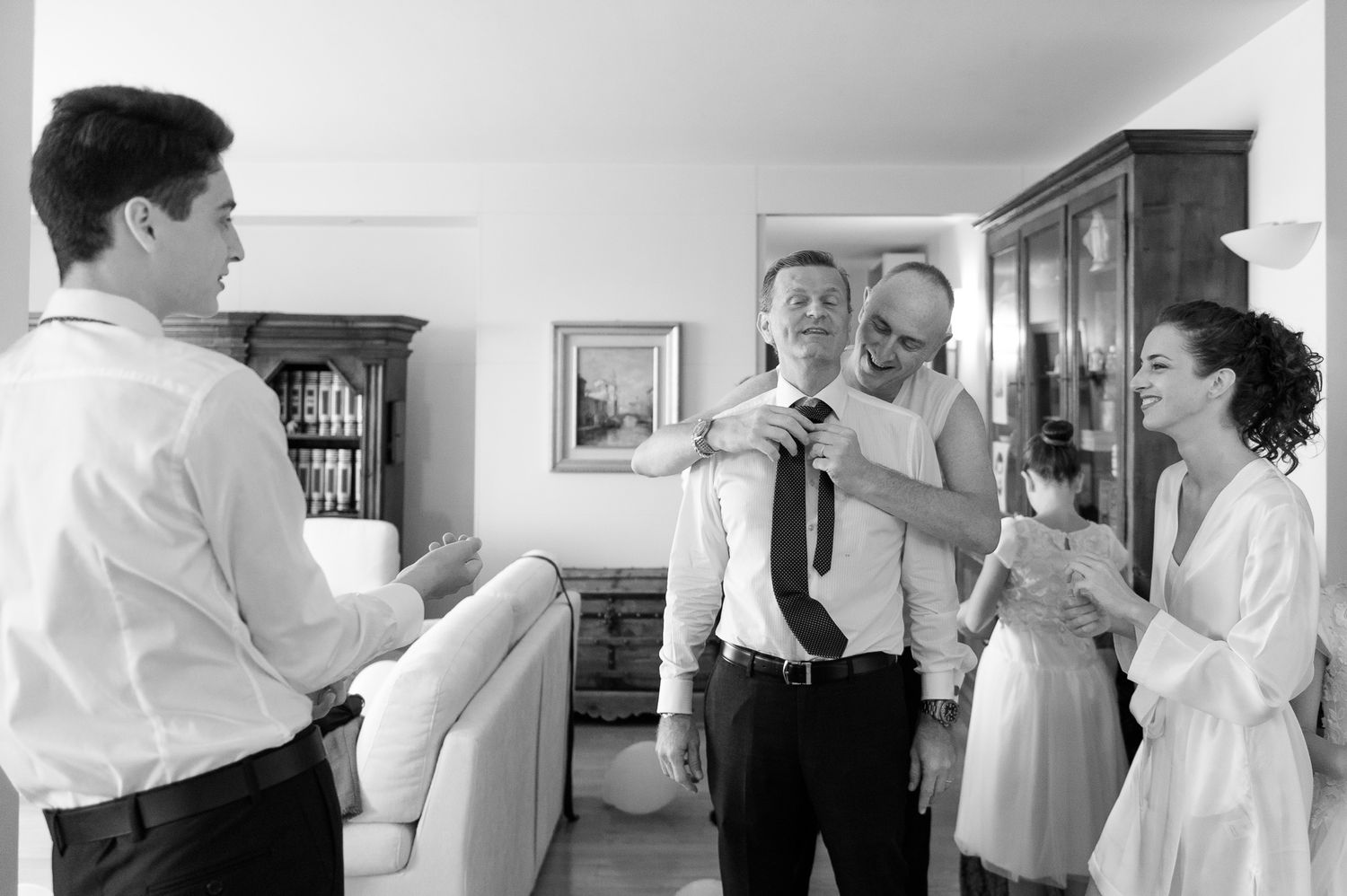 Family helps groom tie his tie while bride watches during wedding preparations.