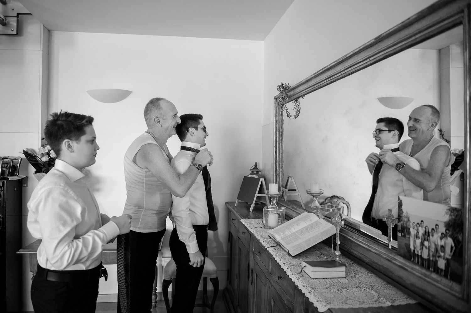Groomsmen assist each other getting dressed before wedding ceremony at mirror.