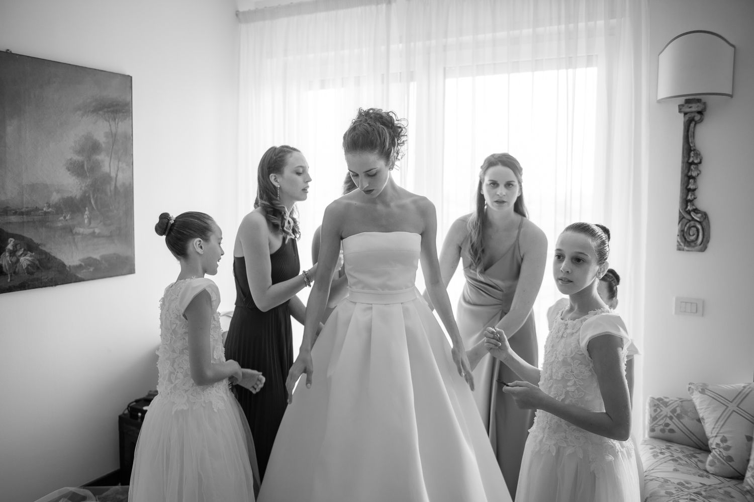 Bride in white gown surrounded by bridesmaids and flower girls helping her prepare in a bright room.