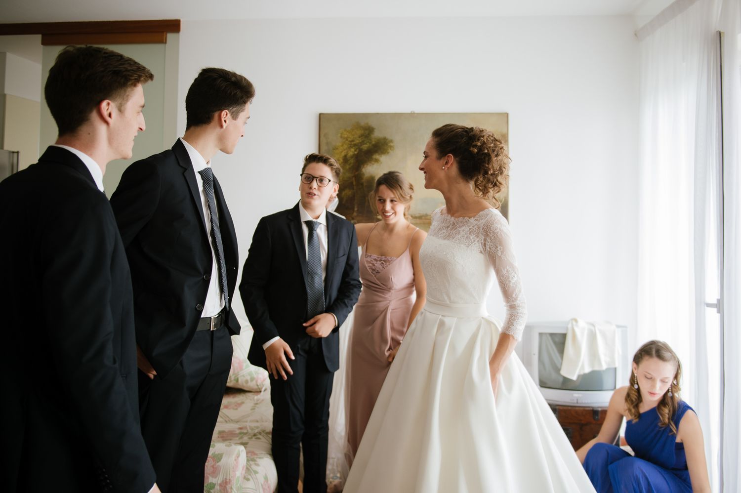 Bride in elegant white gown greeting groomsmen in suits during pre-wedding preparations indoors.