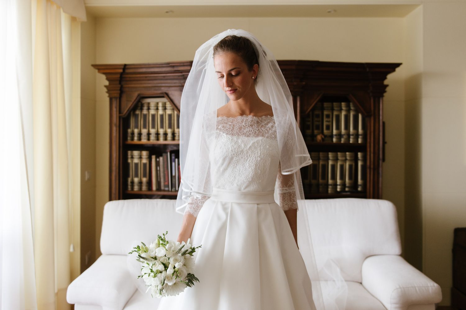 Elegant bride in lace wedding dress and veil holding white bouquet indoors.