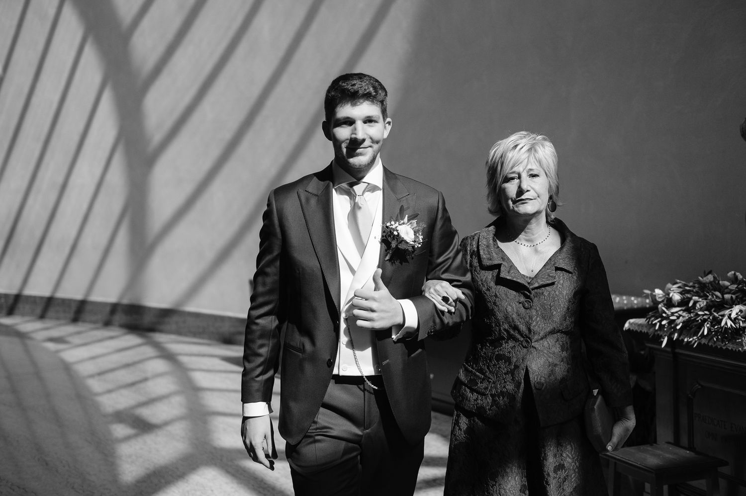 Black and white photo of a smiling groom in a suit walking arm in arm with an older woman.