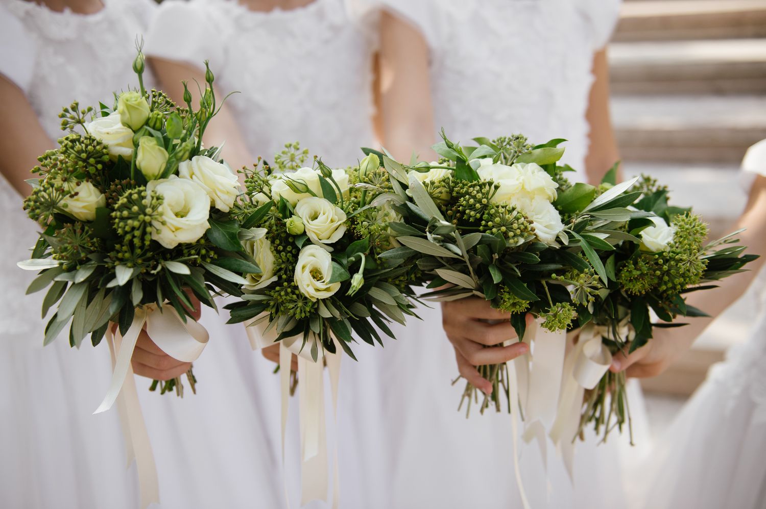Three bridesmaids in white dresses hold matching bouquets of white roses and green blooms with ribbon stems.