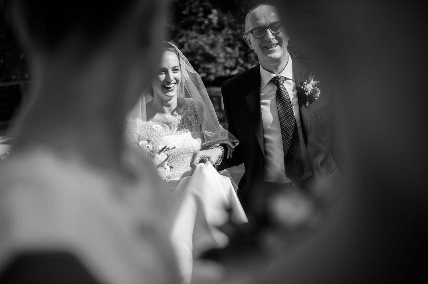 Laughing bride holding bouquet walks with her father in a black and white outdoor wedding photo.