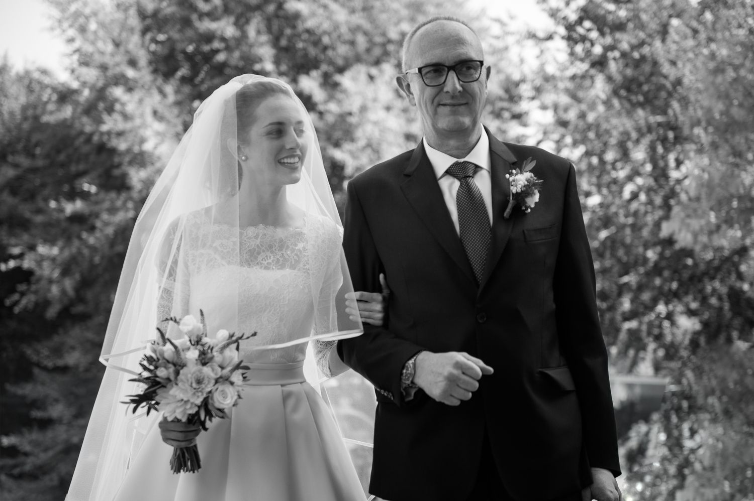 Bride in white gown and veil holding bouquet walks with father in black suit at outdoor ceremony.
