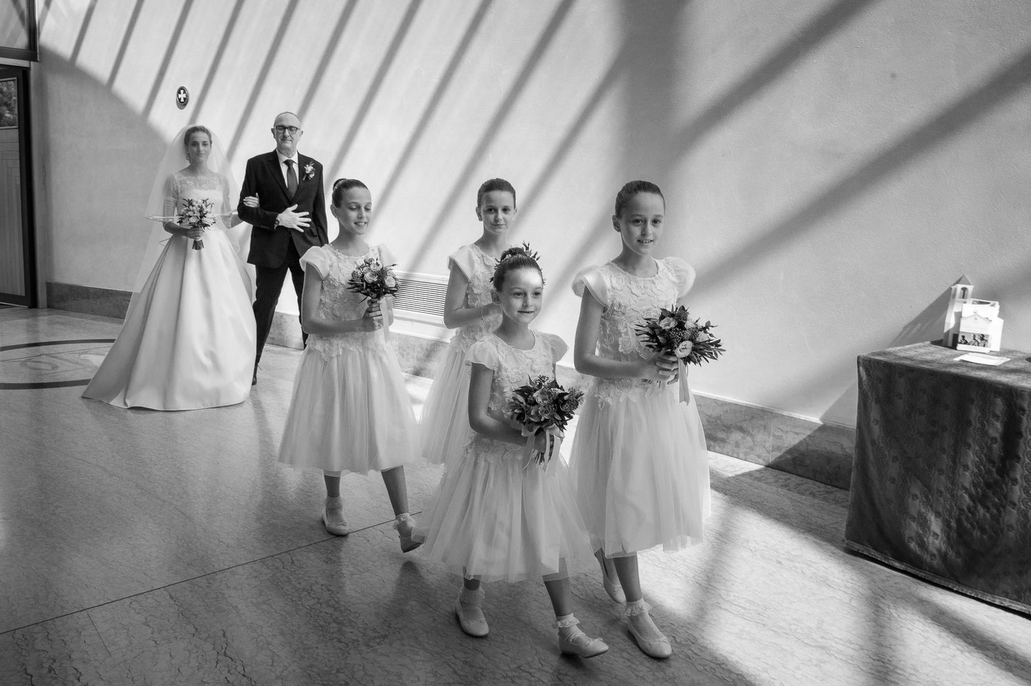 Bride, groom, and flower girls in white dresses posing in sunlit hall before wedding ceremony.