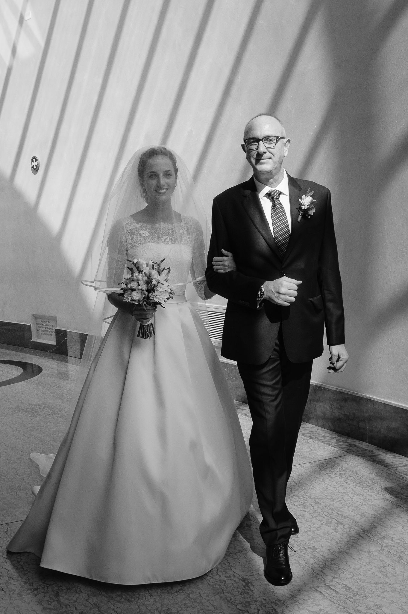Black and white photo of a bride in a lace wedding gown walking down the aisle with her father in a suit.