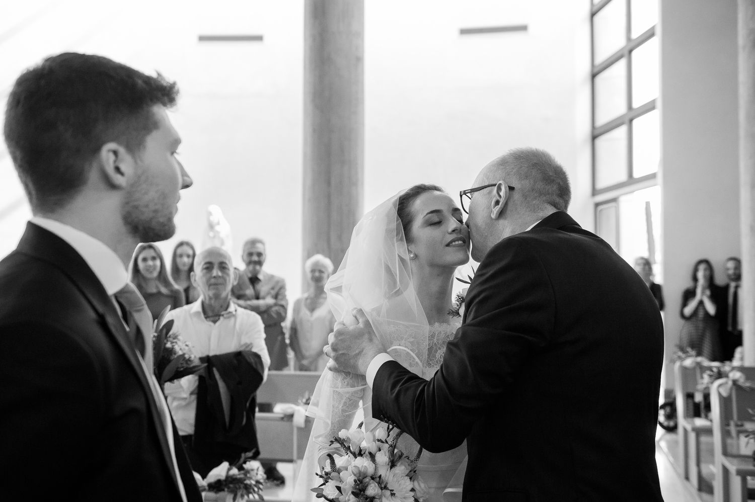 Black and white photo of a bride receiving a kiss from an older man at the church altar.