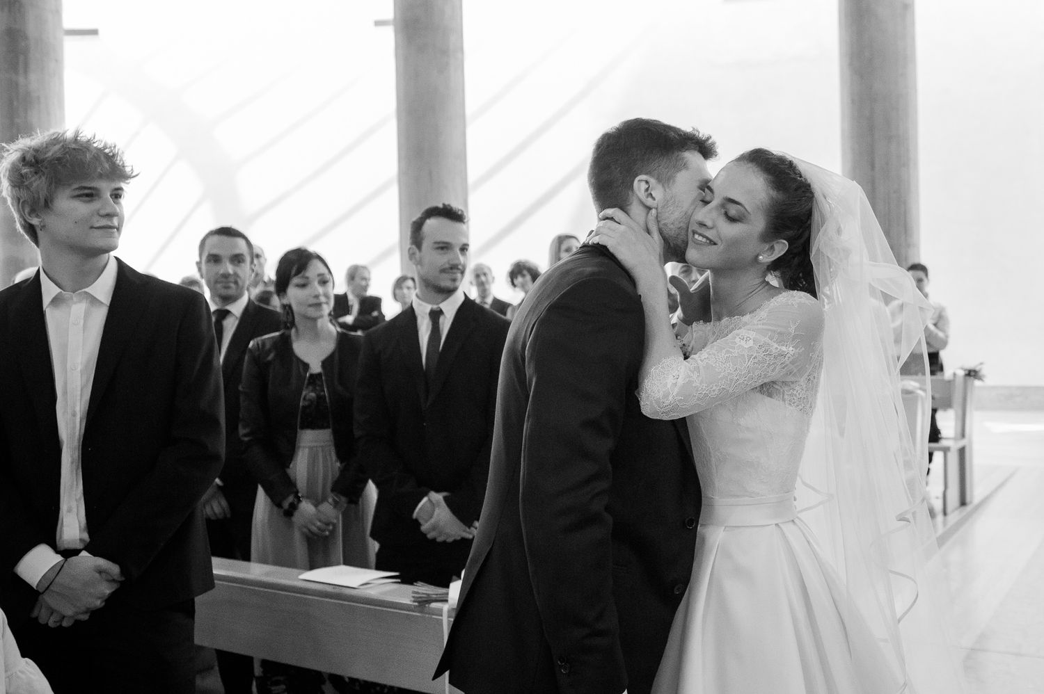 Bride and groom share first kiss at altar during wedding ceremony with guests watching in church.