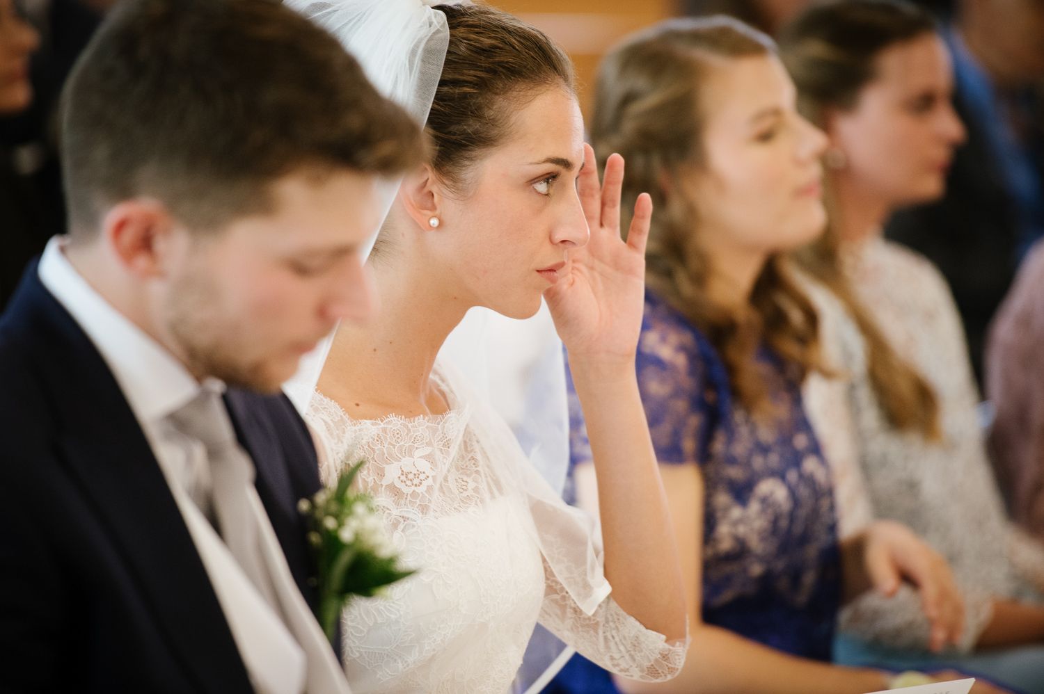 Emotional bride in lace wedding dress wipes tear during church wedding ceremony seated beside groom.