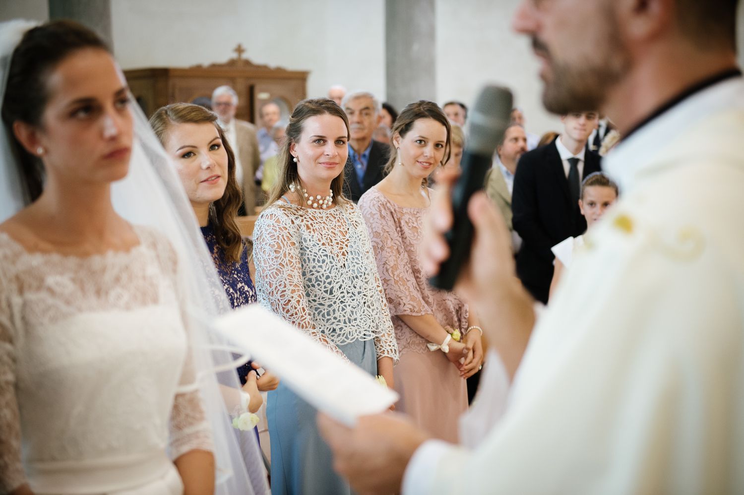 Bride in white lace dress stands with bridesmaids during wedding ceremony in church.