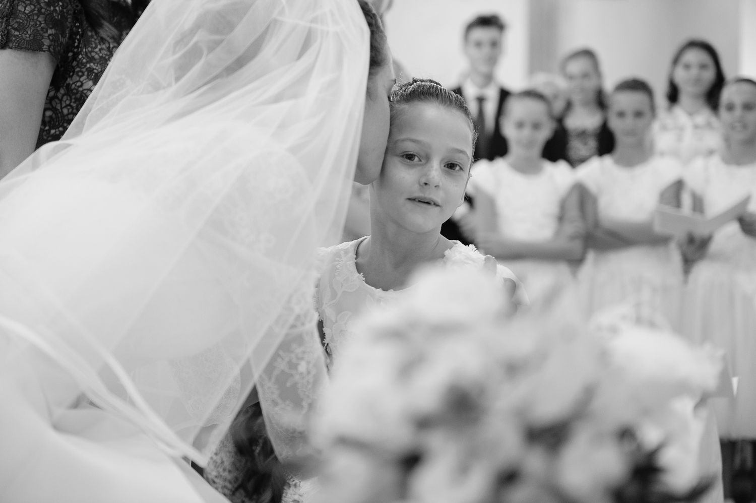 Adorable flower girl in white dress gazing up at the bride during a wedding ceremony, with choir children behind her.