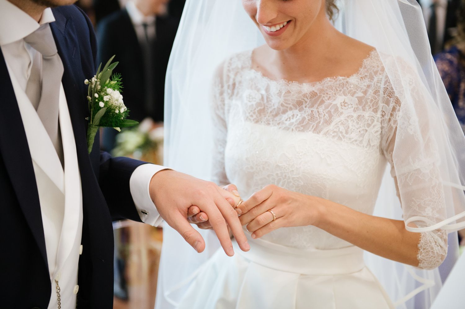 Bride in lace wedding gown smiling as she places a wedding ring on the groom's finger during the ceremony.