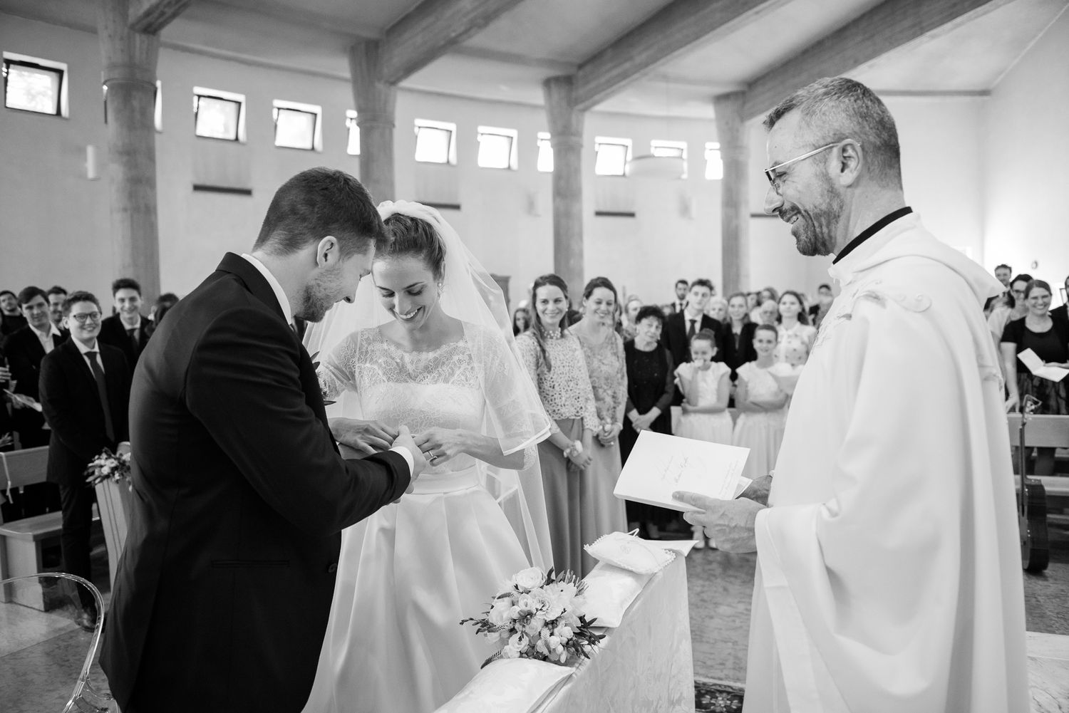 Black and white photo of a bride and groom exchanging rings before a priest during a church ceremony.