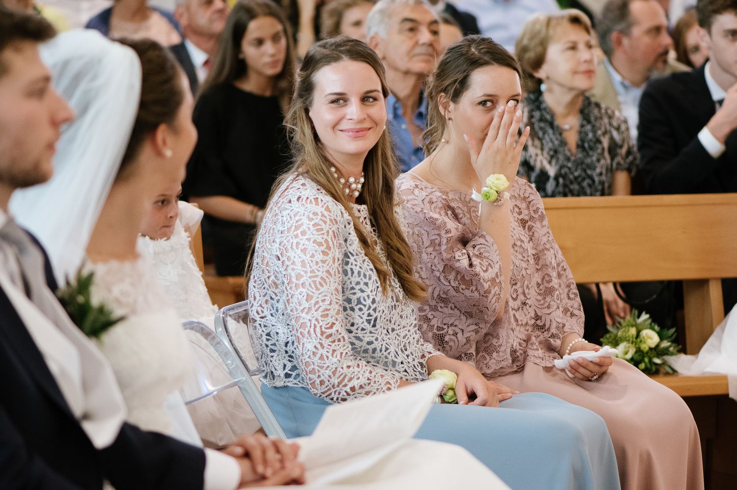 Emotional wedding guests smiling and wiping tears during a church ceremony seated in wooden pews.