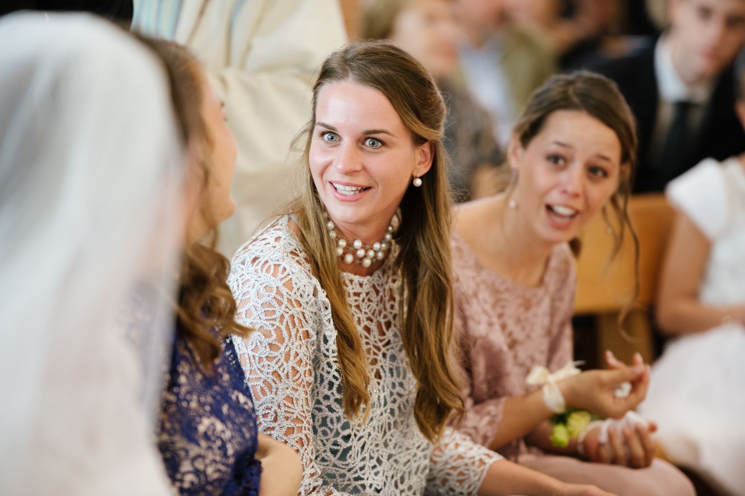 Smiling woman in white lace dress wearing pearl necklace at wedding reception with guests in background.