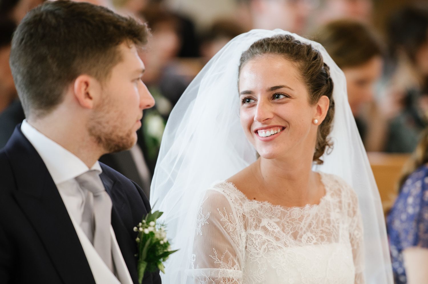 A radiant bride in a lace veil smiles lovingly at her groom during their wedding ceremony.
