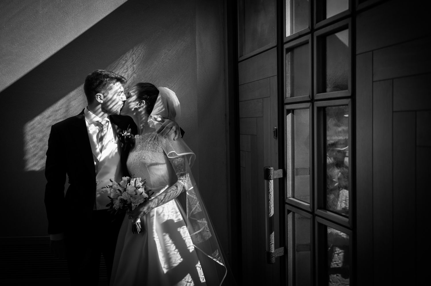 Black and white romantic photo of bride and groom kissing in dramatic light and shadow.