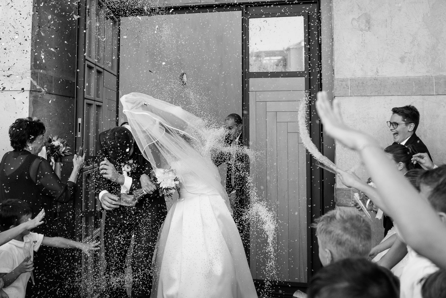 Black and white photo of bride and groom celebrated with confetti outside church doors.