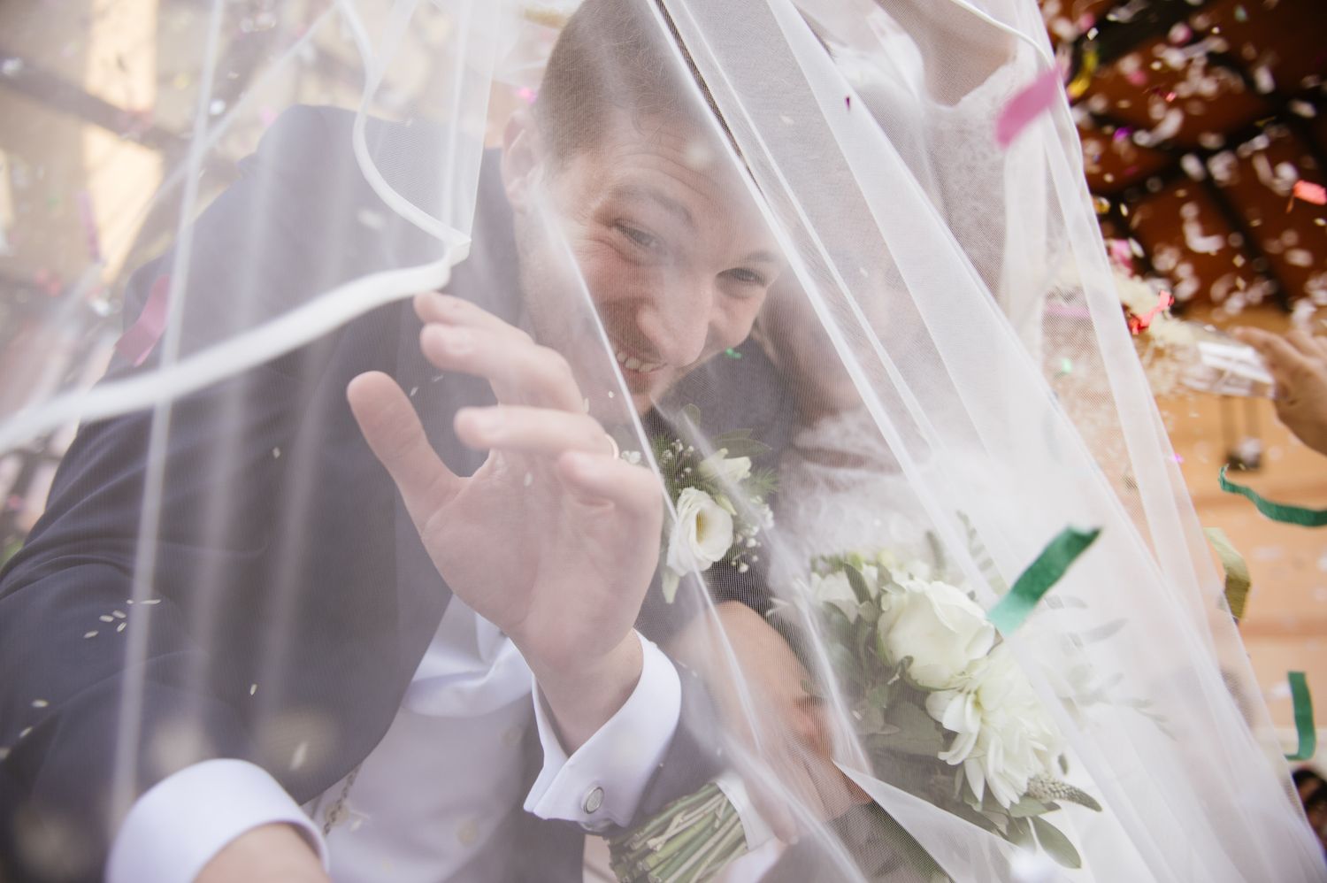Bride and groom sharing intimate moment under wedding veil with confetti and white bouquet.