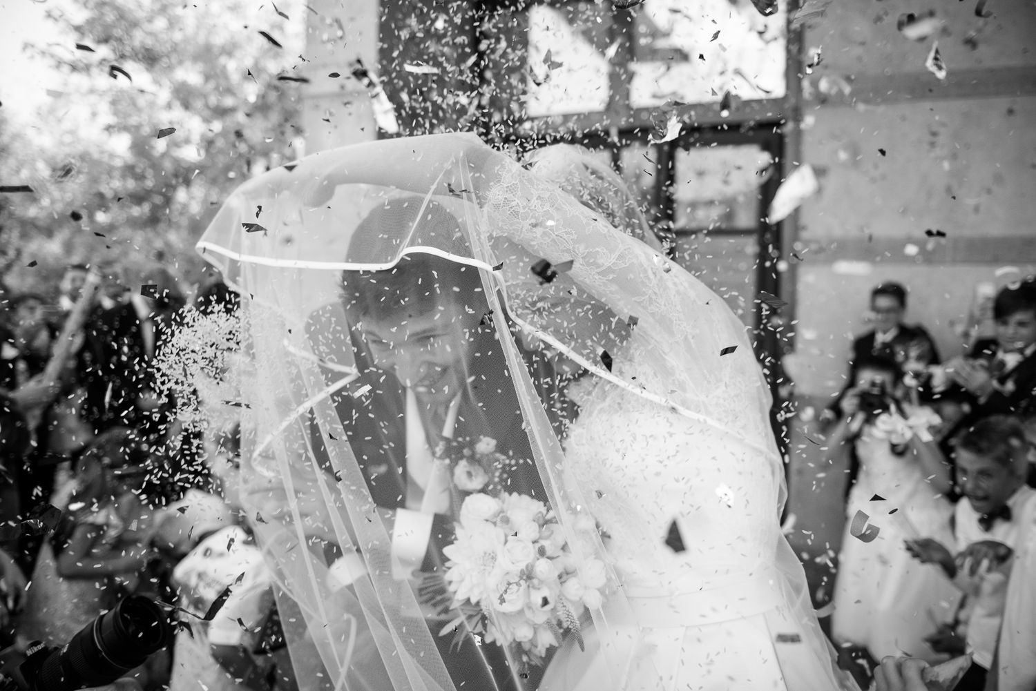 Black and white photo of bride and groom celebrating under veil in shower of confetti.
