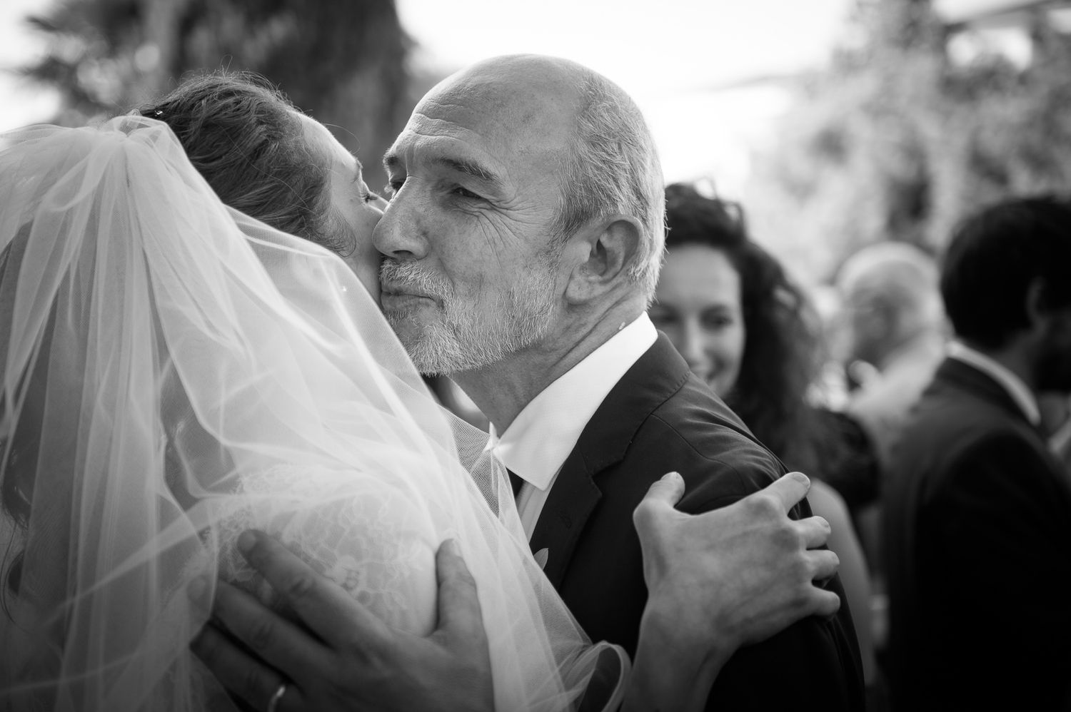 Black and white photo of a bride in veil sharing an emotional hug and kiss with an older man.