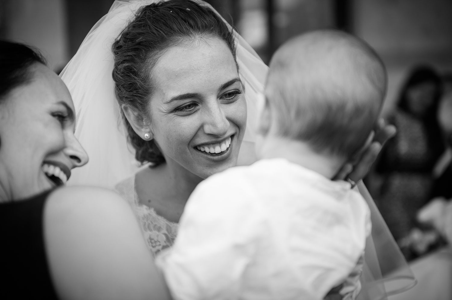 Joyful bride in veil laughing while interacting with baby at wedding reception in black and white.