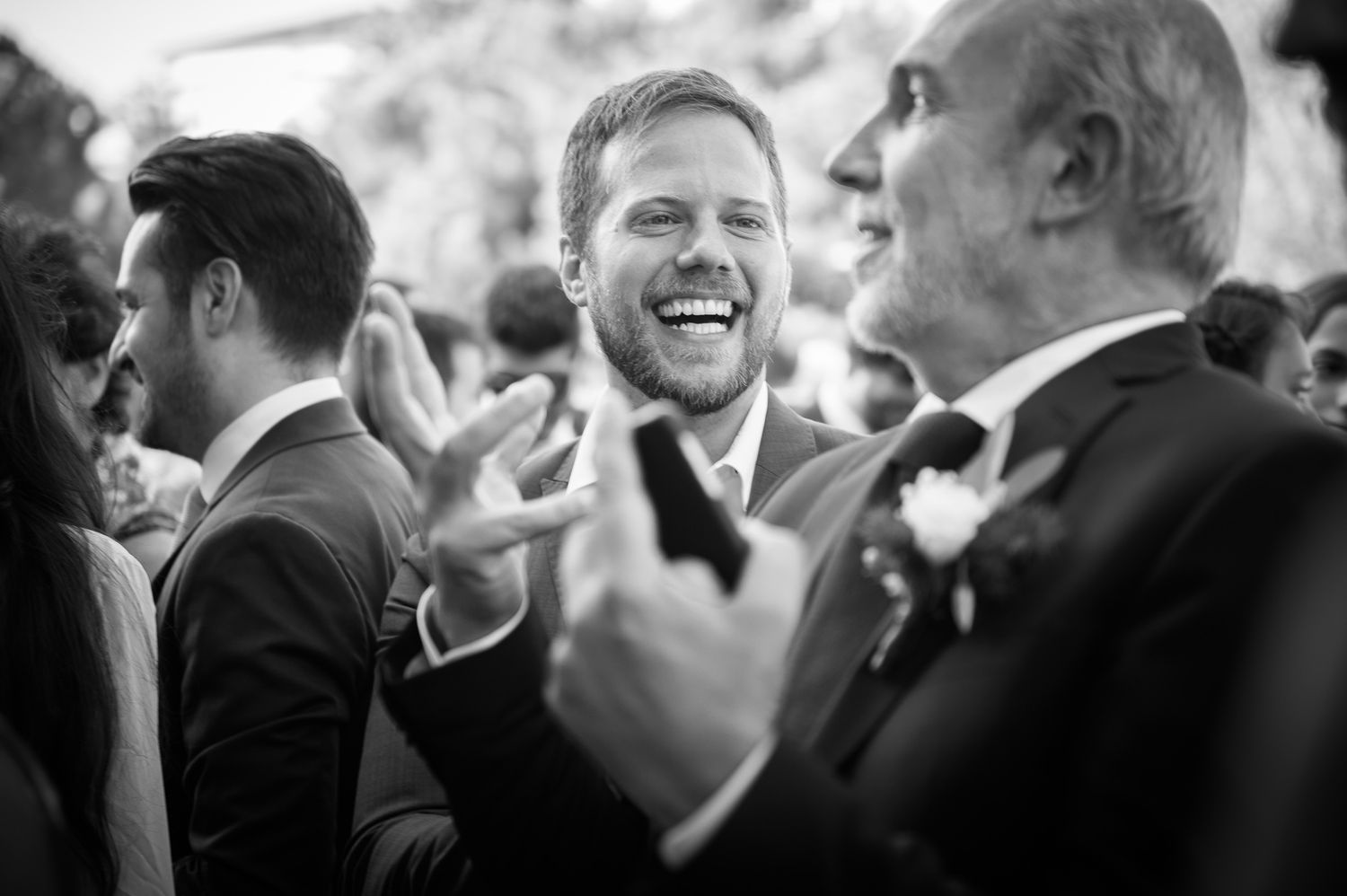 Joyful groom laughing with guests at outdoor wedding reception in black and white photo.