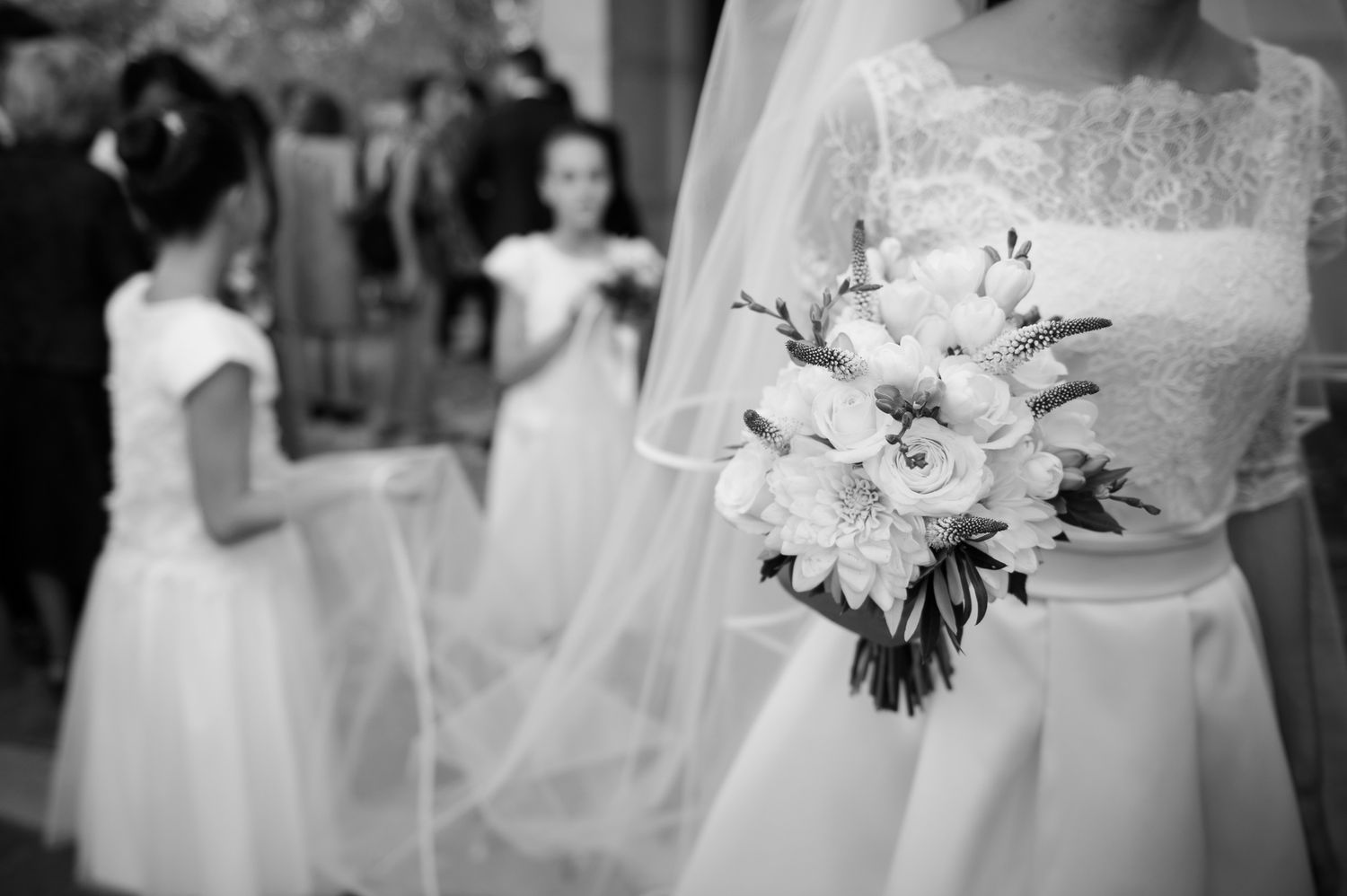 Bride in white lace gown holding bouquet of white roses and flowers with flower girls in background.